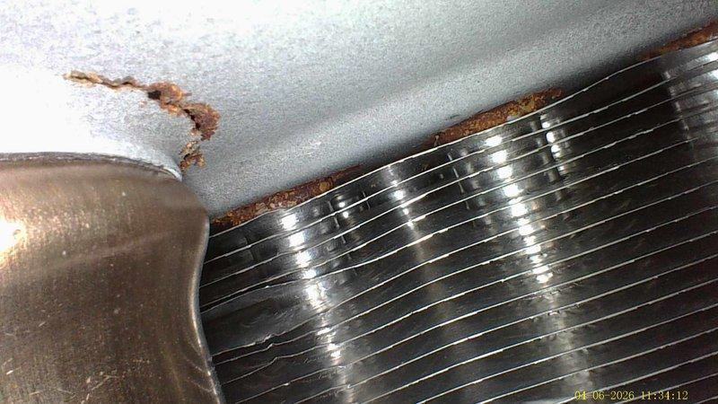 Completed annual cooling maintenance on a 15-year-old air conditioning system. Performed standard tune-up procedures including system inspection, refrigerant line examination, and evaporator coil assessment. Documentation revealed significant rust and corrosion present at the evaporator coil mounting points, with visible deterioration along the top edge where the coil assembly meets the mounting surface. Observed rust-colored staining and water damage on adjacent ceiling surfaces, indicating moisture infiltration or condensation issues. Copper refrigerant lines showed age-appropriate patina and weathering consistent with the system's service life. The finned coil assembly displayed metallic corrosion with rust transfer to surrounding white surfaces, suggesting ongoing moisture exposure that may compromise system efficiency and longevity if left unaddressed.
