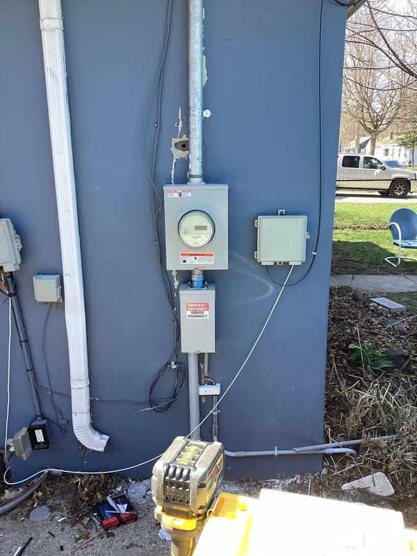 Completed electric service upgrade including coordination with utility company for temporary power disconnect and reconnect. Obtained electrical permit PE26-0355 and scheduled required inspection. Upgraded exterior meter assembly with new gray metal enclosure boxes, circular glass-covered electric meter, and associated junction boxes mounted on building exterior wall. Installed galvanized metal conduit painted white running vertically along wall for proper wire routing. Connected black electrical service wires from meter assembly through conduit system. Passed final electrical inspection and restored power to residence.