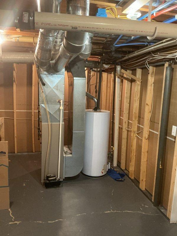Conducted water heater estimate in unfinished basement area with exposed framing and utilities. Assessed existing white cylindrical water heater positioned near HVAC furnace unit, documented current installation conditions including surrounding utility connections, ductwork configuration, and concrete floor placement with existing cracking. Photographed site conditions to provide comprehensive estimate for water heater replacement or service needs.