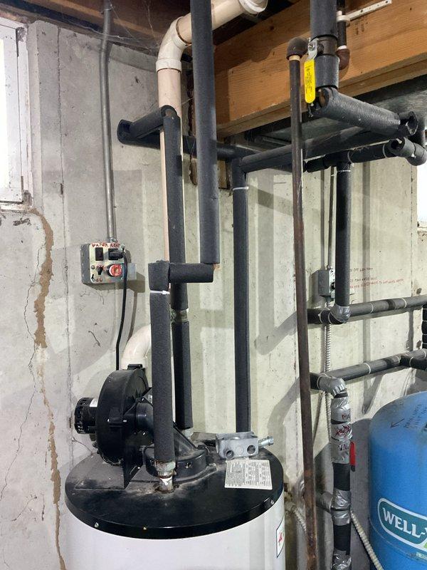 Responded to no hot water call at residential property with approximately 10-year-old water heater system. Upon arrival, assessed basement mechanical room installation including white cylindrical water heater with circulator pump mounted on top, associated piping system with multiple connections, and Well-X-Tec pressure tank. Documented existing system configuration showing complex overhead plumbing arrangement with various T-joints, elbows, and insulated pipe sections routing throughout unfinished basement space. Photographed complete installation for service records.