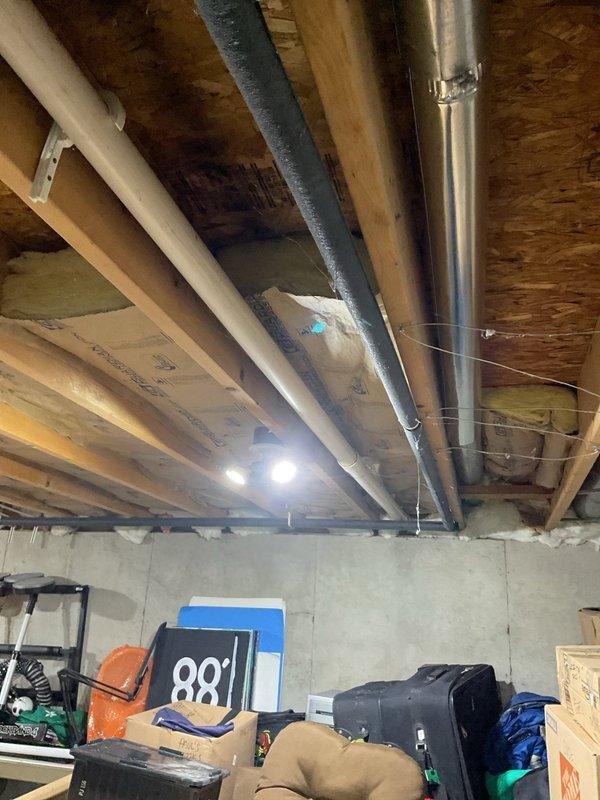 Responded to no hot water call at residential property with approximately 10-year-old water heater system. Upon arrival, assessed basement mechanical room installation including white cylindrical water heater with circulator pump mounted on top, associated piping system with multiple connections, and Well-X-Tec pressure tank. Documented existing system configuration showing complex overhead plumbing arrangement with various T-joints, elbows, and insulated pipe sections routing throughout unfinished basement space. Photographed complete installation for service records.