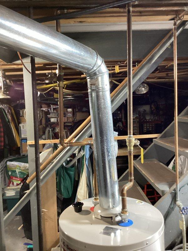 Completed water heater estimate at residential property. Documented existing white cylindrical water heater installation in basement utility area with significant condensation observed on galvanized vent pipe, indicating potential ventilation or humidity concerns. Photographed current plumbing configuration including copper supply lines, brass fittings, shutoff valves, and PVC drainage connections for replacement specifications.