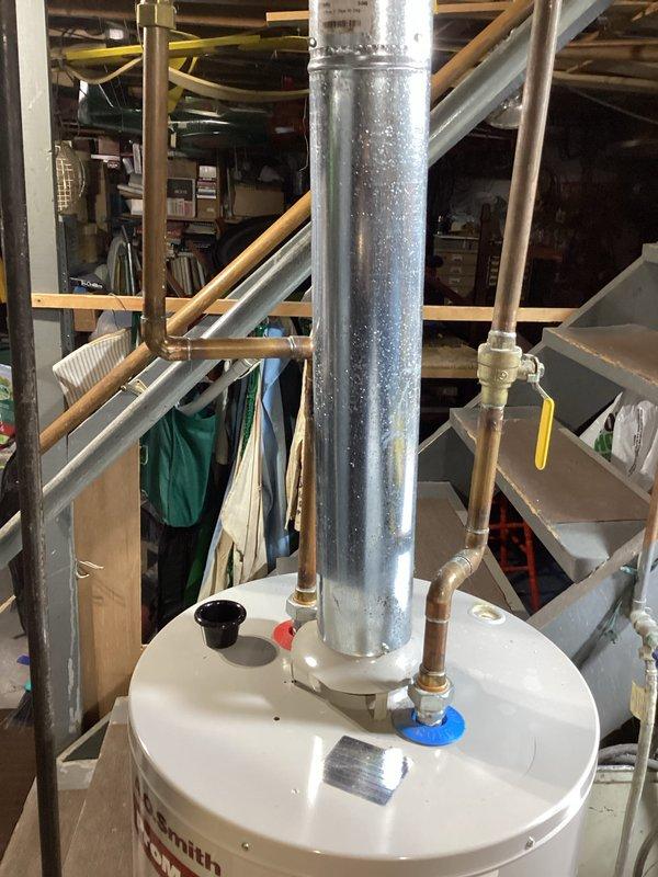 Completed water heater estimate at residential property. Documented existing white cylindrical water heater installation in basement utility area with significant condensation observed on galvanized vent pipe, indicating potential ventilation or humidity concerns. Photographed current plumbing configuration including copper supply lines, brass fittings, shutoff valves, and PVC drainage connections for replacement specifications.