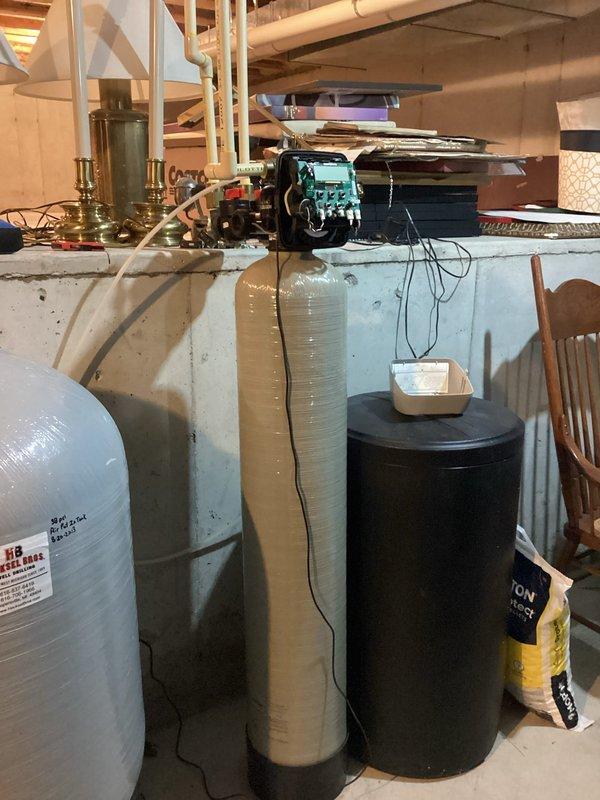 Responded to service call for 19-year-old Stirling water softener reported as not functioning properly. Upon arrival, inspected complete water treatment system including softener tank with tan insulation jacket, brine tank, and associated plumbing components. Evaluated control valve head, copper and PVC supply lines, and brass manifold connections for proper operation and potential failure points. Documented system configuration and current condition with photographic evidence for service records.