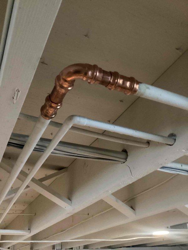 Responded to reported toilet leak in 67-year-old residential property. Upon inspection, identified active leak in overhead plumbing system within basement ceiling area. Leak originated from deteriorated section of waste line requiring immediate repair to prevent further water damage.

Performed repair by removing compromised pipe section and installing new copper piping with compression fittings to bridge connection between existing white PVC drain lines. Configured copper section with appropriate elbow joints and threaded connections to ensure proper alignment and secure integration with existing plumbing infrastructure. All joints were properly fitted and tightened to manufacturer specifications.

Tested repaired section for leaks by running water through the system and verified no active dripping or seepage at connection points. Documented repair work with photographs showing completed copper pipe installation integrated into existing ceiling-mounted plumbing network. Customer advis