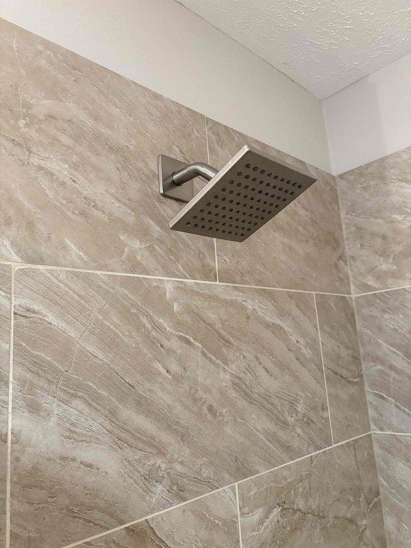 Completed estimate for replacement of 12-year-old residential water heater, with company to supply and install new unit. Assessment included installation of customer-supplied rainfall showerheads in bathroom with marble-effect tile surround. Documented existing bathroom fixtures featuring brushed nickel rectangular rainfall showerhead mounted via angular wall arm, contemporary tile work in beige and taupe tones with natural veining patterns installed in horizontal brick-lay pattern, and two-tone wall paint scheme. Photographed installation areas to document current conditions and fixture specifications for replacement planning purposes.