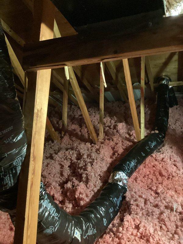 Insulated bathroom exhaust ductwork in attic space to prevent heat loss and condensation issues. Wrapped flexible black exhaust duct with appropriate insulation material and secured metallic silver flexible ducts running through attic joists, ensuring proper support and thermal efficiency throughout the ventilation system.