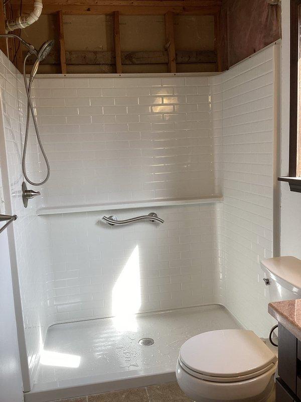 Completed installation of walk-in shower system including white subway tile walls in brick-lay pattern, molded shower pan with center drain, chrome shower head assembly, and horizontal chrome grab bar for safety. All plumbing fixtures installed and operational, with proper water supply connections and drainage completed per code requirements.