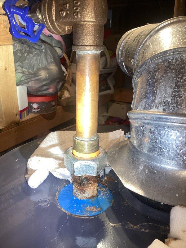 Responded to service call for water heater leak on unit less than 10 years old. Upon inspection, identified active leak at threaded brass fitting connection on cold water inlet pipe. Corrosion and rust evident at hex-shaped coupling, with water staining visible on platform beneath connection point. Blue shutoff valve and copper supply lines showed oxidation and patina consistent with age and moisture exposure. Documented plumbing configuration and leak source with photos showing corroded connection points, condensation on adjacent components, and water damage to surrounding area. Customer advised of findings and recommended repair options for compromised fitting to prevent further water damage to utility space.
