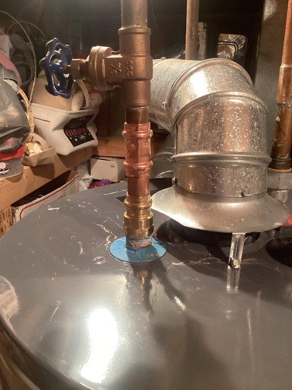 Responded to service call for water heater leak on unit less than 10 years old. Upon inspection, identified active leak at threaded brass fitting connection on cold water inlet pipe. Corrosion and rust evident at hex-shaped coupling, with water staining visible on platform beneath connection point. Blue shutoff valve and copper supply lines showed oxidation and patina consistent with age and moisture exposure. Documented plumbing configuration and leak source with photos showing corroded connection points, condensation on adjacent components, and water damage to surrounding area. Customer advised of findings and recommended repair options for compromised fitting to prevent further water damage to utility space.