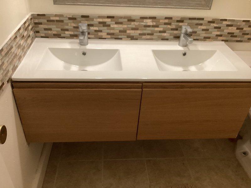 Raised floating double-sink bathroom vanity by 2 inches and reworked the drain line connections to accommodate the new height. The vanity features a white countertop with integrated rectangular basins, wood-grain cabinet drawers, and chrome faucets, all reinstalled and tested for proper function and drainage after the height adjustment was completed.