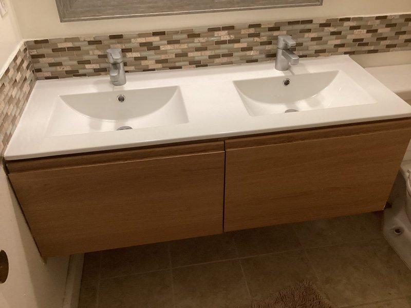 Raised floating double-sink bathroom vanity by 2 inches and reworked the drain line connections to accommodate the new height. The vanity features a white countertop with integrated rectangular basins, wood-grain cabinet drawers, and chrome faucets, all reinstalled and tested for proper function and drainage after the height adjustment was completed.