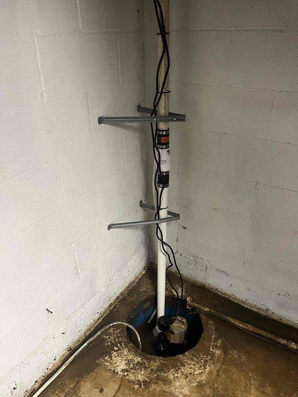 Installed new sump pump system in basement utility area with vertical PVC discharge line secured by metal brackets along corner wall junction. Reworked laundry drain line in crawl space, replacing deteriorated horizontal piping with new white PVC components including elbow fittings and proper mounting hardware to wooden structural supports.