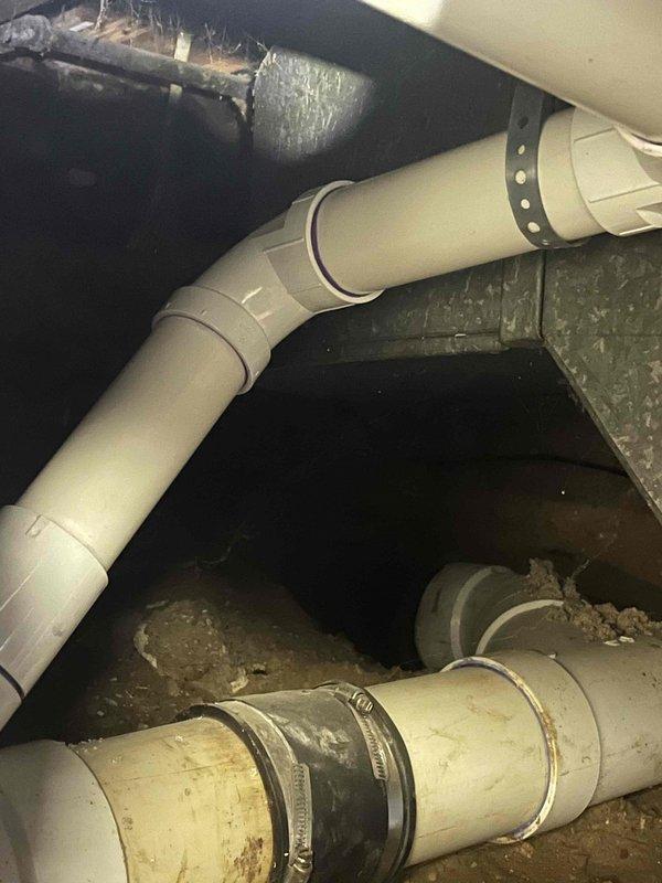 Installed new sump pump system in basement utility area with vertical PVC discharge line secured by metal brackets along corner wall junction. Reworked laundry drain line in crawl space, replacing deteriorated horizontal piping with new white PVC components including elbow fittings and proper mounting hardware to wooden structural supports.