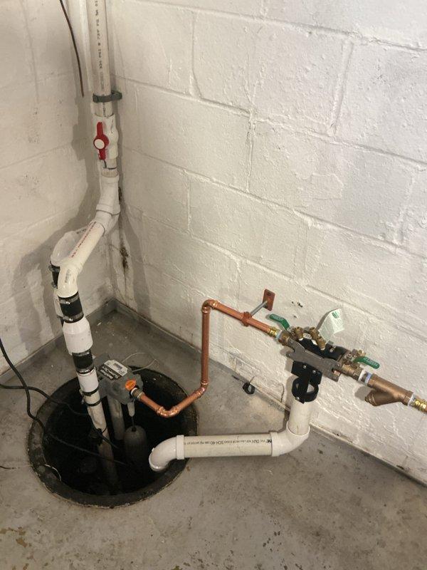 Responded to water backup issue in basement utility area. Upon arrival, assessed existing sump pump system and drainage configuration in corner installation with white cinderblock walls and concrete flooring. Identified compromised drainage capacity causing water accumulation and floor staining.

Installed comprehensive sump pump drainage system with new pump unit positioned in circular pit. Configured vertical discharge piping using white PVC with secure wall mounting straps and integrated red-handled shutoff valve for maintenance access. Established horizontal drainage runs incorporating both PVC and copper piping sections with appropriate transition fittings.

Installed multiple shutoff valves including green-handled ball valve for system isolation and service capability. Connected electrical supply with proper cord routing. Configured secondary drainage pathways with brass fittings and additional valve points to ensure redundant water removal capacity. Mounted all piping securely t
