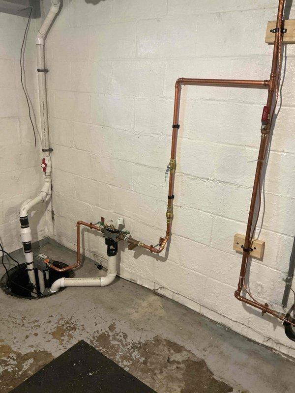 Responded to water backup issue in basement utility area. Upon arrival, assessed existing sump pump system and drainage configuration in corner installation with white cinderblock walls and concrete flooring. Identified compromised drainage capacity causing water accumulation and floor staining.

Installed comprehensive sump pump drainage system with new pump unit positioned in circular pit. Configured vertical discharge piping using white PVC with secure wall mounting straps and integrated red-handled shutoff valve for maintenance access. Established horizontal drainage runs incorporating both PVC and copper piping sections with appropriate transition fittings.

Installed multiple shutoff valves including green-handled ball valve for system isolation and service capability. Connected electrical supply with proper cord routing. Configured secondary drainage pathways with brass fittings and additional valve points to ensure redundant water removal capacity. Mounted all piping securely t