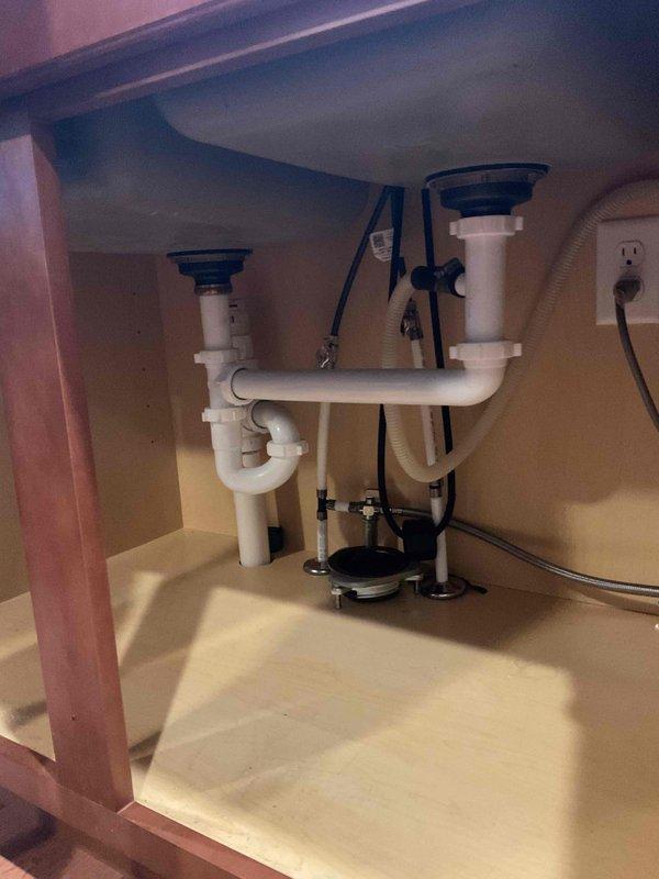 Responded to plumbing lead for garbage disposal installation consultation. Upon arrival, inspected existing under-sink plumbing configuration featuring double basin sink with standard white PVC drain assembly and P-trap system. Evaluated cabinet space, electrical outlet availability, and existing plumbing connections to assess feasibility for garbage disposal installation. Documented current setup showing dual drain tailpieces, P-traps, and adequate electrical service present in cabinet. Provided client with assessment of installation requirements and discussed disposal unit mounting options for the existing sink configuration.
