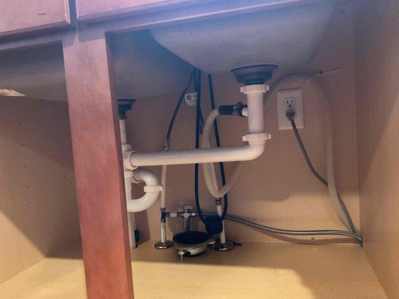 Responded to plumbing lead for garbage disposal installation consultation. Upon arrival, inspected existing under-sink plumbing configuration featuring double basin sink with standard white PVC drain assembly and P-trap system. Evaluated cabinet space, electrical outlet availability, and existing plumbing connections to assess feasibility for garbage disposal installation. Documented current setup showing dual drain tailpieces, P-traps, and adequate electrical service present in cabinet. Provided client with assessment of installation requirements and discussed disposal unit mounting options for the existing sink configuration.