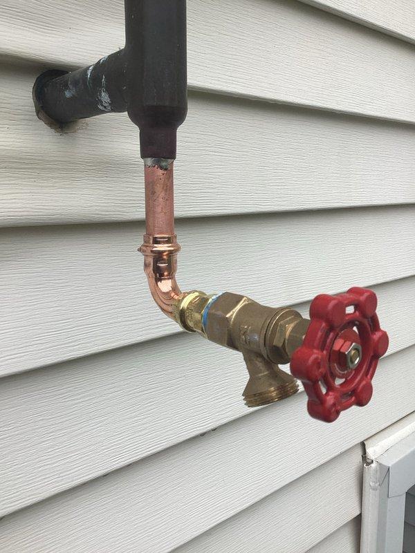 Responded to service call regarding sprinkler supply line ball valve that was not replaced during previous visit. Upon inspection, found portion of sprinkler water supply system disconnected but confirmed no active leaking present. Documented existing conditions showing weathered vertical black pipe connected to oxidized copper piping with brass valve assembly featuring red wheel-style gate valve handle. System components displayed typical outdoor weathering with visible corrosion at connection points and patination on copper sections. Photographed current installation mounted on exterior siding for reference and communicated findings to team for customer follow-up as requested.