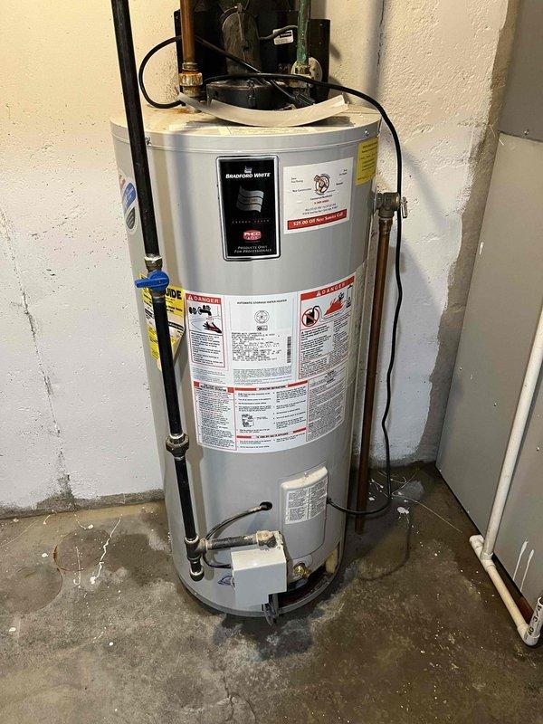 Completed estimate for replacement of 20-year-old Bradford White residential water heater located in basement utility area. Unit was installed in 2006 and has been drained by homeowner in preparation for replacement. Existing installation includes standard plumbing connections with copper supply lines, gas line with brass fittings, blue-handled shutoff valve, drainage pan, and circulator pump assembly mounted on top of unit. Documented current configuration and measurements for replacement specifications.