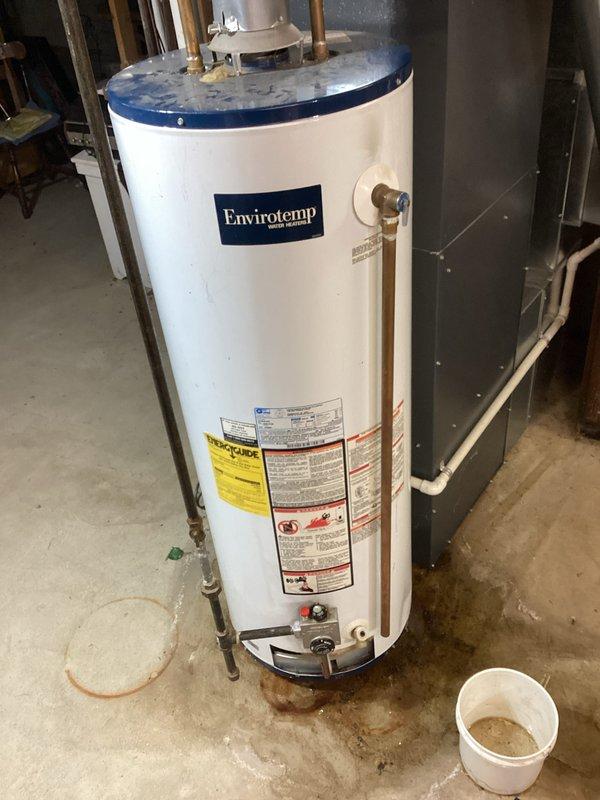 Responded to service call for leaking Envirotemp water heater with water accumulation present at base of unit for approximately three to four days. Upon inspection, confirmed active leak underneath water heater with visible water staining on concrete floor surrounding unit. Documented equipment condition with photographs showing copper plumbing connections, unit specifications, and extent of water damage in utility area.