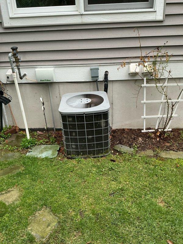 Conducted free estimate for residential air conditioning system replacement. Existing outdoor condenser unit is 22 years old, barrel-style configuration with cylindrical design and mesh cage protection, located on concrete pad in side yard landscaping area. Indoor equipment includes Tempstar furnace with Aprilaire humidifier in basement utility room. Documented current system configuration with photos for replacement proposal and equipment sizing recommendations.