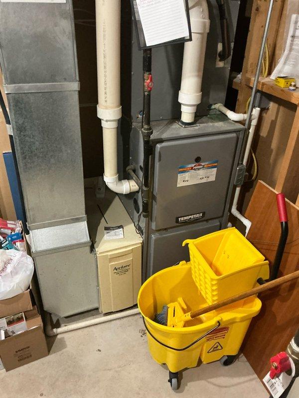 Conducted free estimate for residential air conditioning system replacement. Existing outdoor condenser unit is 22 years old, barrel-style configuration with cylindrical design and mesh cage protection, located on concrete pad in side yard landscaping area. Indoor equipment includes Tempstar furnace with Aprilaire humidifier in basement utility room. Documented current system configuration with photos for replacement proposal and equipment sizing recommendations.