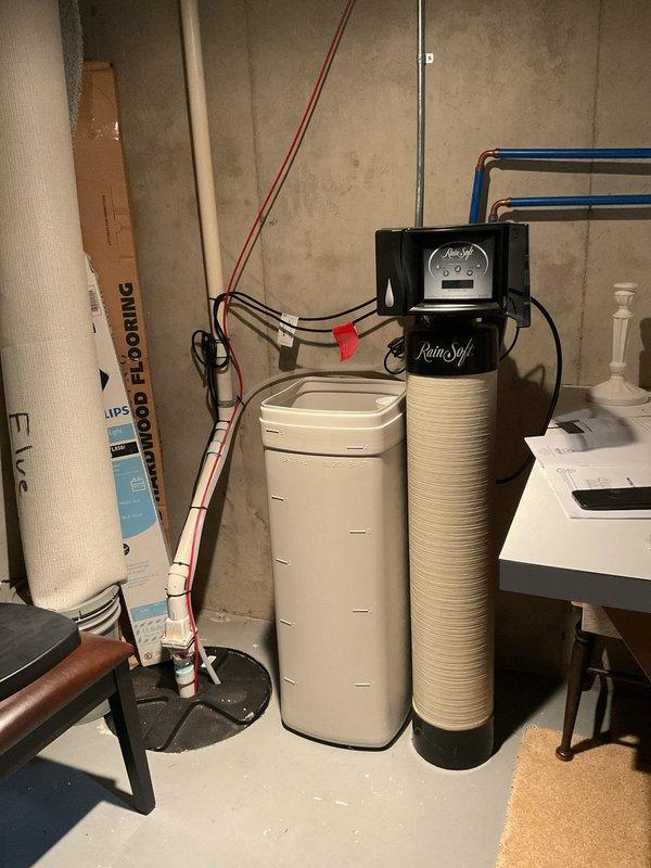 Responded to service call for RainSoft water softener and purification system verification in 20-year-old home where functionality concerns existed for past 3 years. Inspected dual-tank water treatment system in basement, including brine tank and resin tank with control head, confirmed proper plumbing connections via copper and PEX piping throughout house. Verified salt level in brine tank appeared adequate with visible salt pellets present for regeneration cycle.