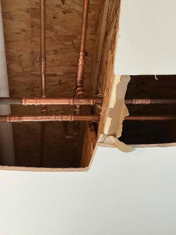Responded to report of water spotting on second-floor ceiling with suspected leak from upstairs bathrooms. Cut two access openings in ceiling to investigate moisture trapped between drywall and existing popcorn ceiling layers. Installed temporary plastic containment barriers secured with painter's tape at both openings to prevent debris contamination. Exposed and inspected copper plumbing lines running through ceiling cavity, identifying multiple pipe runs servicing two full bathrooms above. Documented conditions with photographs showing plumbing configuration and ceiling structure for further assessment and repair planning.