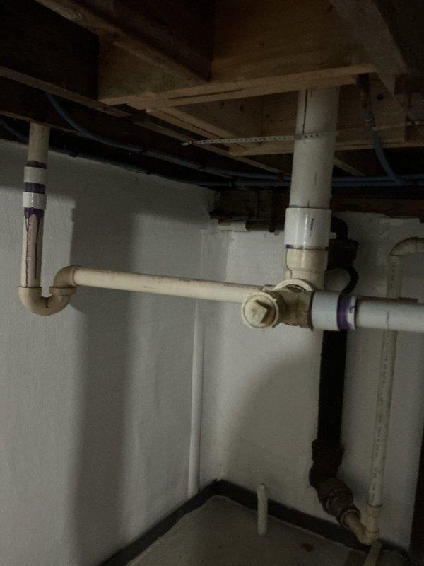 Responded to plumbing leak in basement beneath bathroom fixture in 1940s-era single family home. Upon inspection, identified leak originating from PVC drain assembly connected to bathroom sink, which had been actively leaking for several weeks. Accessed plumbing system from basement level where exposed wooden joists and drain piping were visible. Examined white PVC P-trap configuration and associated drain lines, noting purple primer markings at connection points. Assessed horizontal and vertical pipe runs, including connections between white PVC drainage system and black ABS waste lines. Inspected all joints, fittings, and couplings in the drain assembly for signs of failure or improper installation. Documented existing plumbing configuration including older metal piping connected to newer plastic drainage system. Repaired leak at affected connection point, ensuring proper seal and drainage flow. Verified repair eliminated active water intrusion and confirmed system functionality befo