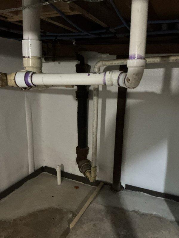 Responded to plumbing leak in basement beneath bathroom fixture in 1940s-era single family home. Upon inspection, identified leak originating from PVC drain assembly connected to bathroom sink, which had been actively leaking for several weeks. Accessed plumbing system from basement level where exposed wooden joists and drain piping were visible. Examined white PVC P-trap configuration and associated drain lines, noting purple primer markings at connection points. Assessed horizontal and vertical pipe runs, including connections between white PVC drainage system and black ABS waste lines. Inspected all joints, fittings, and couplings in the drain assembly for signs of failure or improper installation. Documented existing plumbing configuration including older metal piping connected to newer plastic drainage system. Repaired leak at affected connection point, ensuring proper seal and drainage flow. Verified repair eliminated active water intrusion and confirmed system functionality befo