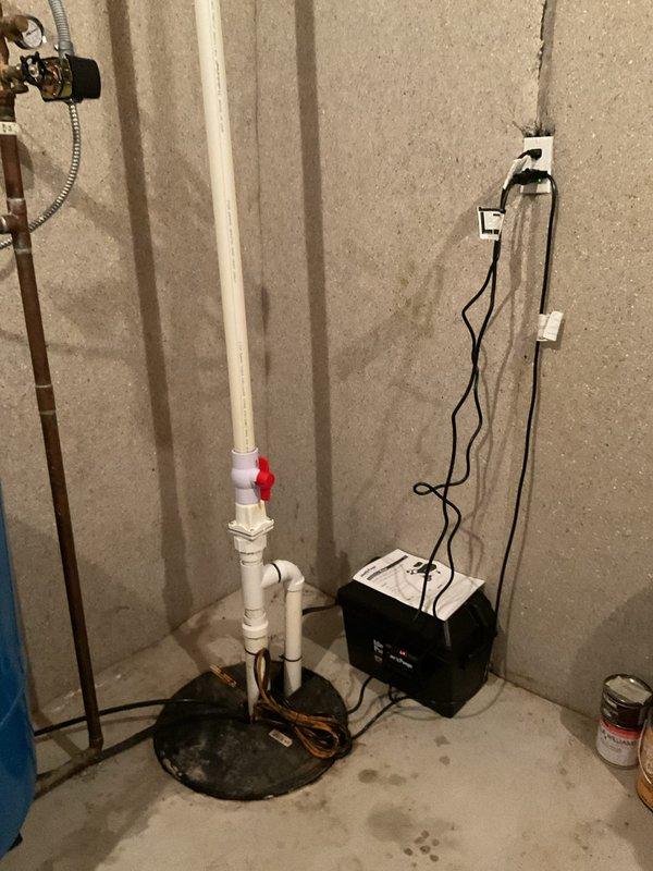 Installed battery backup system for existing sump pump in basement utility area. Connected backup power supply unit to sump pump basin assembly with proper electrical wiring secured along concrete block wall, ensuring system will maintain operation during power outages. Verified all PVC discharge piping connections and tested system functionality upon completion.