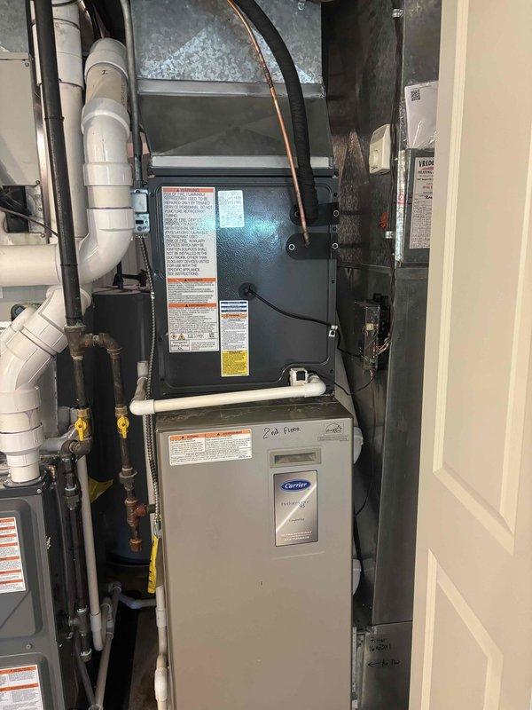 Completed full HVAC system installation including indoor air handler and Carrier furnace unit mounted vertically in mechanical closet with proper refrigerant line connections, PVC condensate drainage, and ductwork integration. Installed outdoor condenser unit on gravel pad against exterior wall with secure mounting, proper electrical conduit routing, and refrigerant line connections to house. All equipment tested and operational, with manufacturer labels visible and installation meeting code requirements for residential HVAC systems.