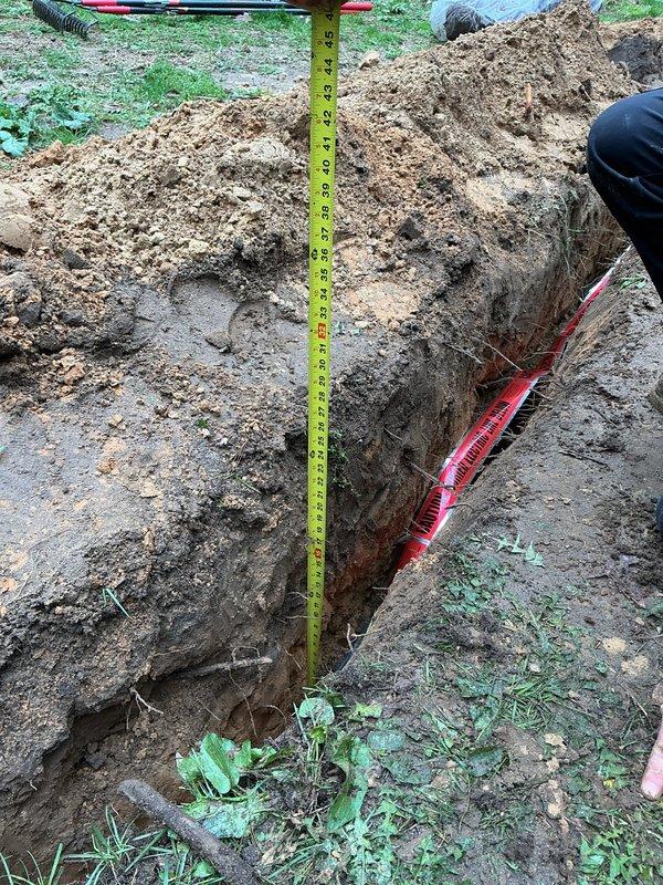 Excavated trench to access and install red flexible conduit piping from outbuilding to main structure. Trench depth measured and verified, conduit laid and secured along trench bottom, with excavated soil staged for backfill upon completion.