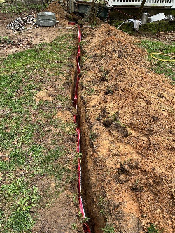 Excavated trench to access and install red flexible conduit piping from outbuilding to main structure. Trench depth measured and verified, conduit laid and secured along trench bottom, with excavated soil staged for backfill upon completion.