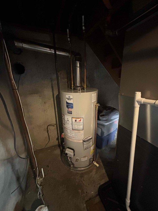 Completed water heater installation in basement utility area. Installed new white cylindrical water heater unit with proper metallic exhaust vent piping extending to ceiling for safe combustion gas ventilation. Connected all necessary plumbing lines including hot and cold water supply lines with appropriate valves and fittings at base of unit. Secured all copper piping connections and verified proper clearances around unit for safety and code compliance. Ensured all manufacturer specifications and regulatory requirements were met during installation. Tested unit operation and confirmed proper venting system function. Work area left clean and operational.