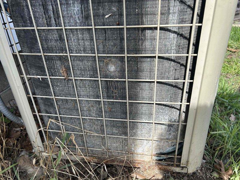 # Cooling System Maintenance and Assessment

Responded to service call for inadequate cooling performance on a 25-year-old residential air conditioning system. Upon arrival, performed comprehensive visual inspection of outdoor condenser unit, which revealed extensive contamination of condenser coils with accumulated dirt, dust, and debris causing significant airflow restriction. Heavy buildup was evident throughout coil surfaces, with black discoloration indicating years of neglect. Lower coil sections displayed white crystalline deposits and mineral accumulation, further compromising heat transfer efficiency.

Documented unit condition through photographic evidence showing deteriorated state of condenser coils, overgrown vegetation encroaching on unit perimeter, and weathered protective wire guard. Ground-level debris and organic matter had accumulated around base of unit, contributing to restricted airflow and reduced system performance. The severity of contamination and age of equip