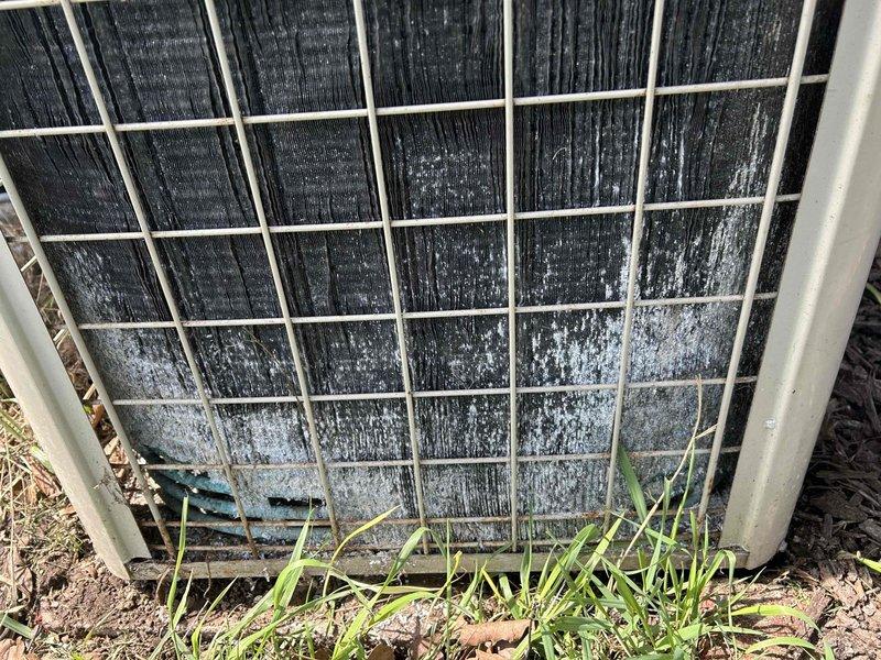 # Cooling System Maintenance and Assessment

Responded to service call for inadequate cooling performance on a 25-year-old residential air conditioning system. Upon arrival, performed comprehensive visual inspection of outdoor condenser unit, which revealed extensive contamination of condenser coils with accumulated dirt, dust, and debris causing significant airflow restriction. Heavy buildup was evident throughout coil surfaces, with black discoloration indicating years of neglect. Lower coil sections displayed white crystalline deposits and mineral accumulation, further compromising heat transfer efficiency.

Documented unit condition through photographic evidence showing deteriorated state of condenser coils, overgrown vegetation encroaching on unit perimeter, and weathered protective wire guard. Ground-level debris and organic matter had accumulated around base of unit, contributing to restricted airflow and reduced system performance. The severity of contamination and age of equip