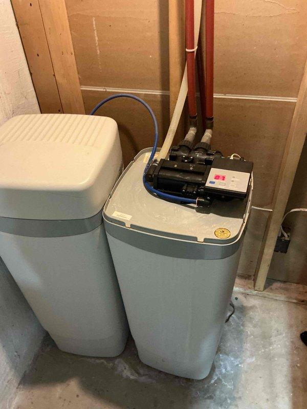 Responded to a water softener leak that had been active for approximately 90 days in a 1969-built home. Upon inspection of the basement utility area, identified the leak source at the water softener plumbing connections. The system consisted of dual water treatment units with copper supply lines and PEX distribution piping mounted on a plywood backing panel. Assessed the vertical piping assembly including shut-off valves, fittings, and connections to determine the exact point of water intrusion. Documented the condition of both the water softener unit and associated brine tank with pump assembly, noting moisture staining on the concrete floor at the base of the units. Photographed the complete plumbing configuration including copper pipes, flexible tubing connections, and control valves for service records. Completed necessary repairs to eliminate the leak and restore proper system operation.