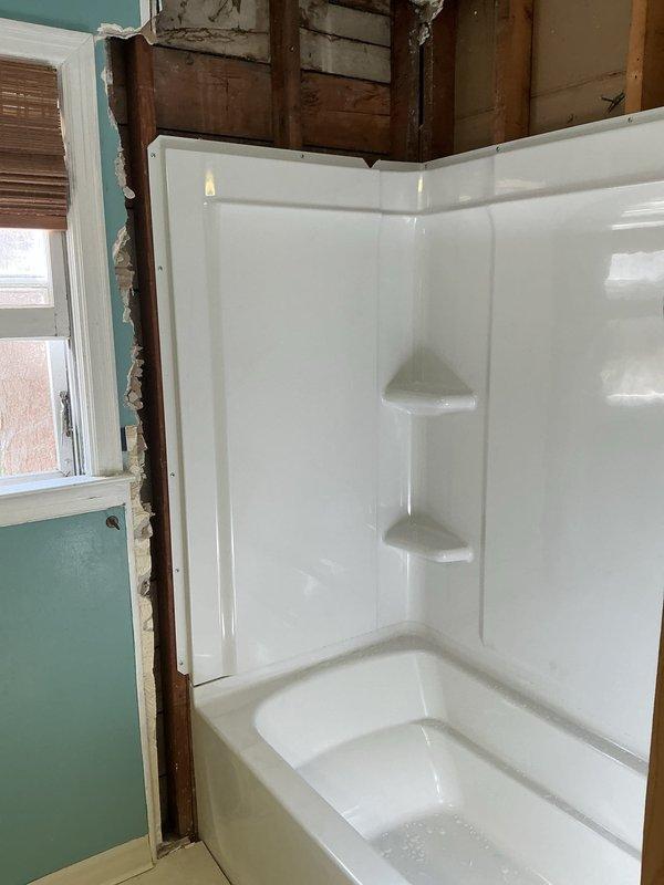 Completed full shower surround replacement in bathroom. Removed existing deteriorated wall materials and fixtures, exposing framing and studs. Installed new white molded fiberglass bathtub and shower surround unit with integrated corner shelving.