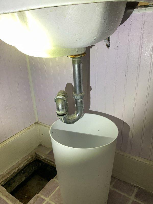 Responded to a second-floor bathroom sink leak complaint. Upon inspection, discovered heavily corroded vertical drain pipe with significant green oxidation and deterioration beneath the sink. The existing P-trap and drainage system showed signs of long-term water damage and corrosion. Assessed the full extent of pipe degradation from the sink connection down through the floor opening. Documented the temporary drainage setup with collection bucket due to compromised pipe integrity. Recommended complete replacement of corroded drainage pipes and P-trap assembly to restore proper function and prevent further water damage to surrounding floor structure and subflooring.