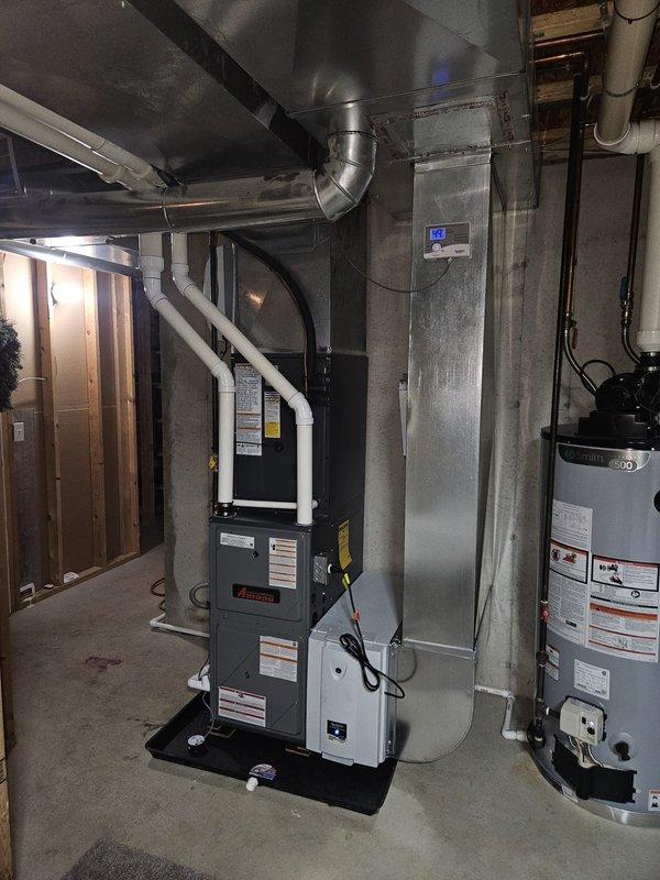 Completed full HVAC system installation including new Amana air conditioning condenser unit and furnace. Mounted outdoor AC unit on concrete pad against exterior wall with proper clearances, installed refrigerant line sets with gray and white conduit runs up vinyl siding to electrical disconnect box. Interior installation included new gas furnace in basement mechanical room with complete ductwork connections to existing distribution system. Installed white PVC venting from furnace through ceiling joists, connected all supply and return ductwork using rectangular and cylindrical metal ducts. Verified proper electrical connections, tested system operation for heating and cooling functions, confirmed all safety controls and limit switches functioning correctly. Placed protective mat under furnace, ensured all manufacturer labels and warning stickers properly affixed to equipment. System commissioned and left in full operational status.