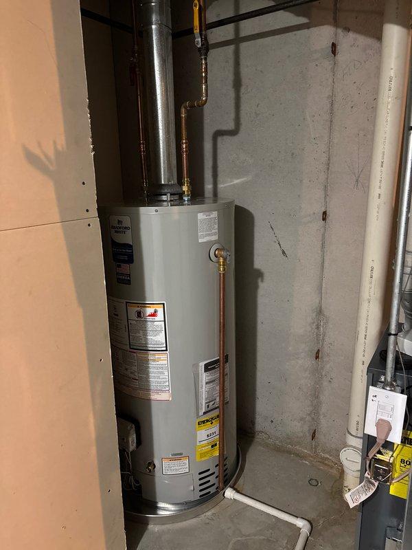 Completed installation of residential water heater in basement utility area. Installed new white cylindrical water heater unit against concrete wall with proper copper piping connections for hot and cold water lines, brass fittings, and metallic vent pipe extending upward. Secured unit with yellow safety restraint chain per code requirements, verified all manufacturer labels and Energy Guide documentation are visible and intact, and ensured proper clearances from electrical panel and adjacent PVC plumbing. Unit is operational and ready for service.
