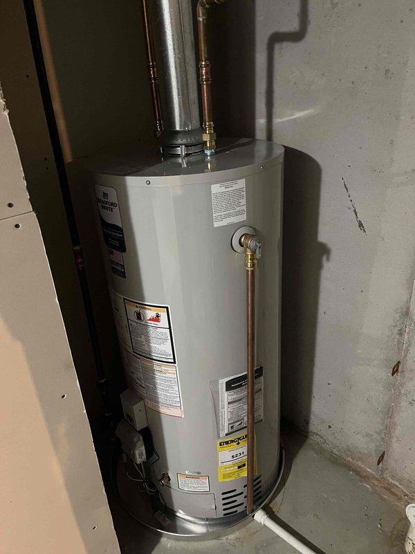 Completed installation of residential water heater in basement utility area. Installed new white cylindrical water heater unit against concrete wall with proper copper piping connections for hot and cold water lines, brass fittings, and metallic vent pipe extending upward. Secured unit with yellow safety restraint chain per code requirements, verified all manufacturer labels and Energy Guide documentation are visible and intact, and ensured proper clearances from electrical panel and adjacent PVC plumbing. Unit is operational and ready for service.