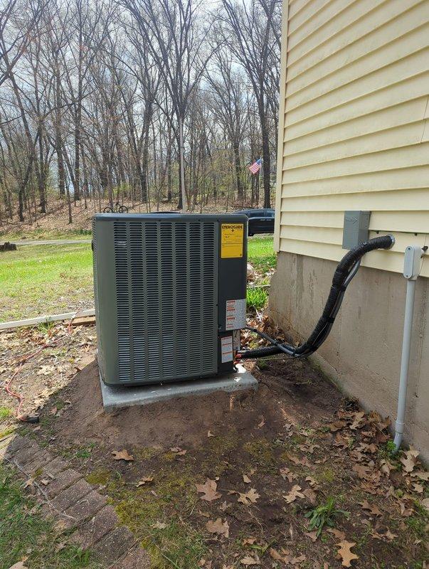 Completed full HVAC system installation including new furnace and air conditioning unit. Interior installation features dark metal plenum chamber with metallic ductwork distribution system, curved and straight duct sections properly sealed and fastened, mounted on concrete foundation with white PVC condensate drainage piping installed along vertical plenum. Exterior condensing unit positioned on concrete pad against house foundation with black insulated refrigerant line set routed through siding to indoor equipment. All ductwork connections secured with proper fasteners and sealing, system installed per manufacturer specifications and ready for operation.