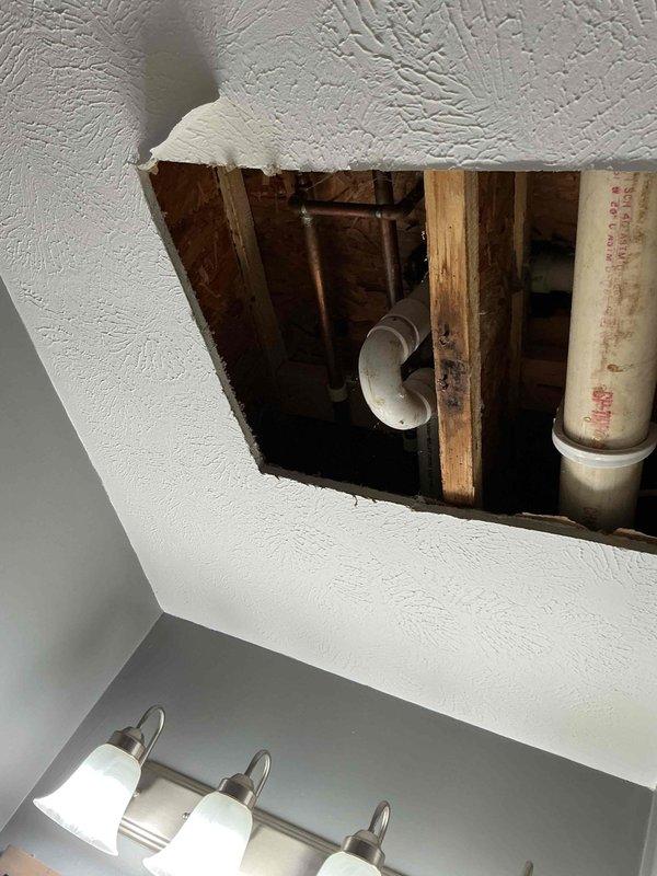 Responded to plumbing leak reported from upstairs floor with water intrusion visible below. Created ceiling access opening to expose and inspect plumbing infrastructure above bathroom vanity area. Located source of leak in overhead piping system, including PVC and metal supply lines. Documented conditions through photographs showing exposed framing, pipe configuration, and leak origin point for repair assessment and future reference.