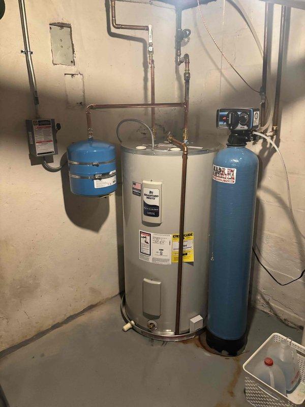 Successfully installed a 50-gallon Bradford White electric water heater (Model RE350S6-1NCWH) with 4500W/3500W dual heating elements in basement utility area. Installation included integration with existing water treatment system consisting of blue pressure tank and water softener/filtration unit, connected via copper piping with appropriate fittings and valves. Added domestic hot water expansion tank to system per code requirements to protect against thermal expansion and maintain proper system pressure.