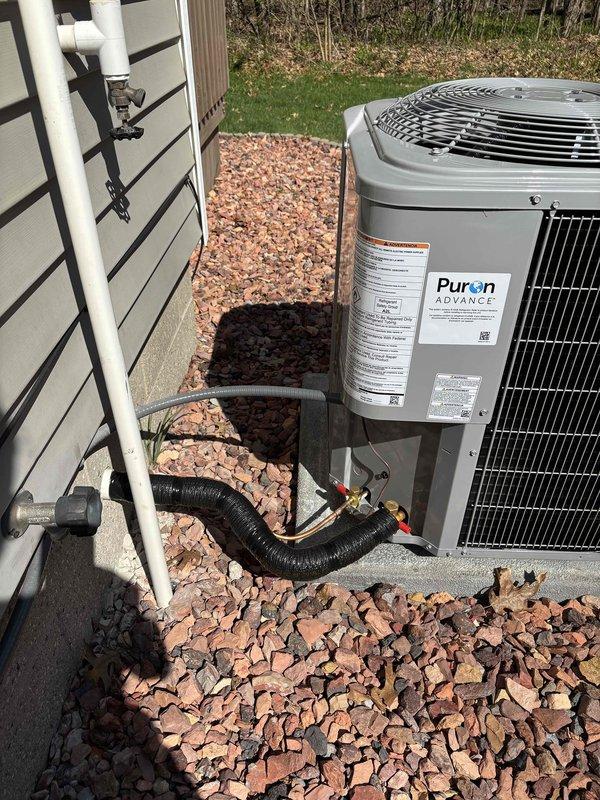 Completed add-on air conditioning installation featuring a Puron ADVANCE outdoor condensing unit mounted on concrete pad with decorative rock landscaping. Installed refrigerant line set with proper insulation, ran electrical conduit through exterior wall penetration with appropriate bushing, and connected condensate drain line. System professionally commissioned with all refrigerant lines secured and protected per manufacturer specifications.