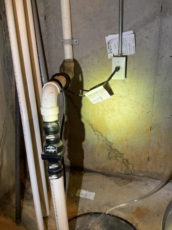 Responded to water heater with extinguished pilot light and no hot water production. Upon arrival, discovered significant water infiltration in basement utility area with standing water on floor surrounding Bradford White water heater installed January 2027. Assessed basement flooding conditions and determined water intrusion was affecting heater operation and posed safety concerns for pilot light system. Documented extensive water damage with yellowish discoloration on concrete floor and walls, along with active sump pump system attempting to manage water levels. Photographed Rockjaw backup sump pump control system with dual activation switches and connected pump assembly, as well as overall flooding conditions affecting the water heater location. Due to unsafe operating conditions created by standing water and compromised pilot light, recommended addressing basement water infiltration issues before attempting pilot relight to prevent recurring outages and ensure safe operation of gas