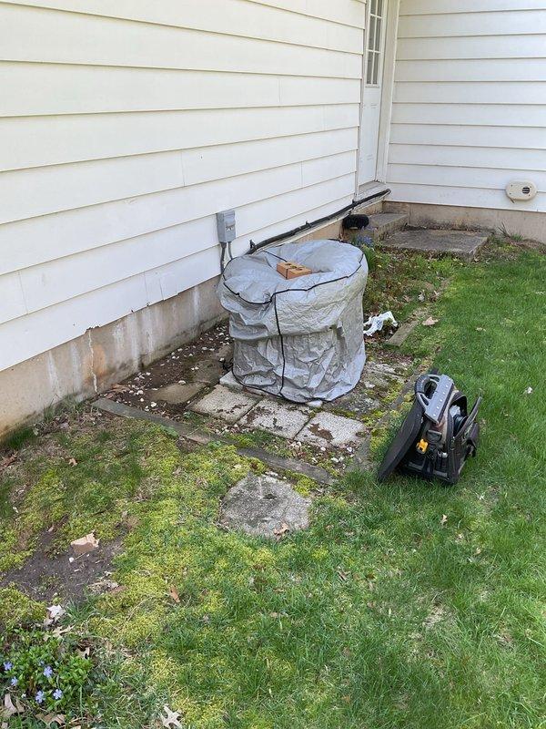 Responded to no-cooling complaint on 20-year-old Carrier air conditioning system. Upon arrival, performed visual inspection of outdoor condensing unit and captured documentation photos showing unit condition with weathered concrete pad and algae growth around equipment area. Condenser fan grille inspection revealed rust-colored discoloration and dirt accumulation around central motor hub, though grille structure remained intact. Unit was blowing air but failing to provide adequate cooling to residence. Documented equipment condition including exterior housing, fan assembly, and surrounding service area for diagnostic evaluation.